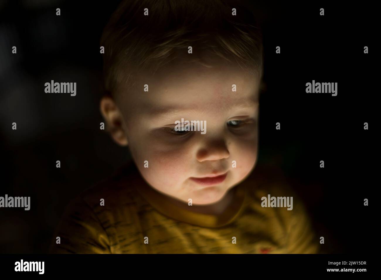 Blonde haired, blue eyed toddler boy looks down with a light shining upwards. Stock Photo