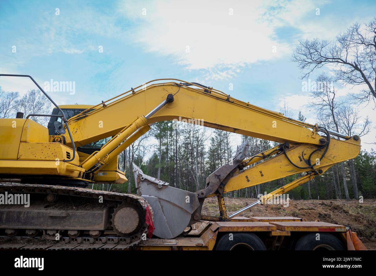 Boom and Bucket of a yellow Skid Steer Excavator Loader on a flat bed trailer, at a construction job site, in evening light with trees in background. Stock Photo