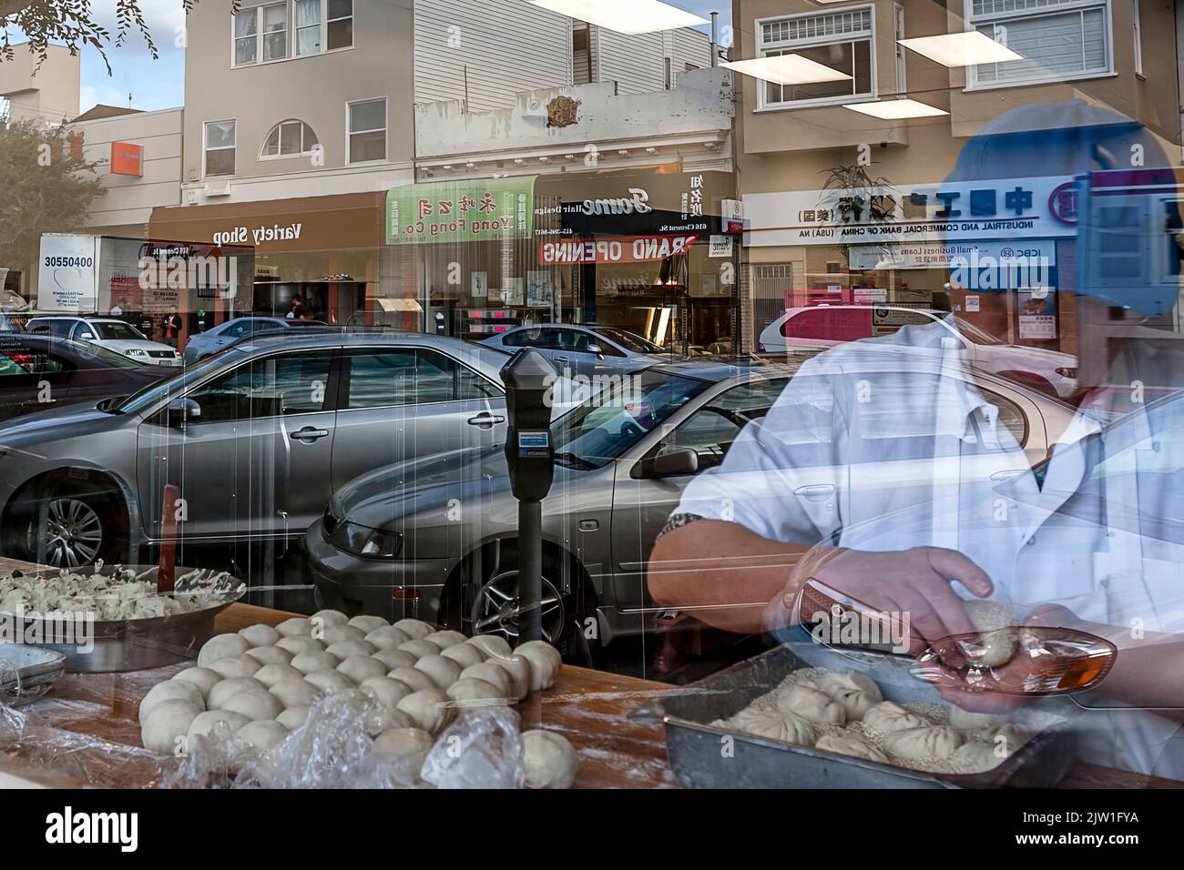 A baker works in his store window with reflections of the street and diagonal parking of the cars and other stores across the street. Stock Photo