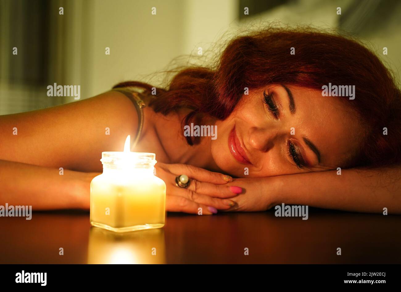 Happy relaxed woman enjoying the aroma of candle. Smiling girl looks at a burn candle in home cosiness in the evening. Rest self care Stock Photo