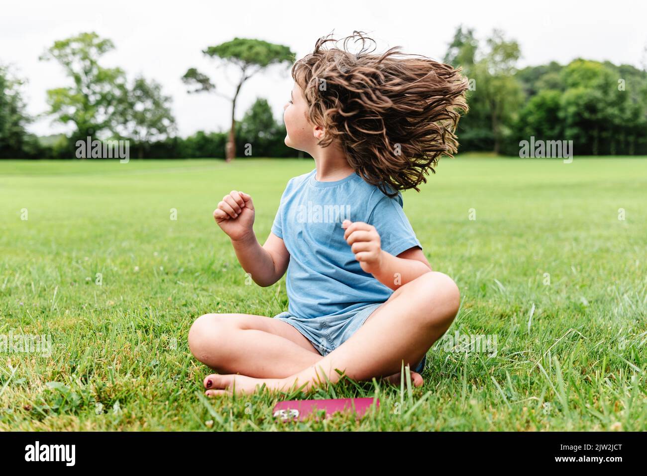 Cheerful barefoot child in casual clothes smiling and shaking hair while sitting cross legged on grassy lawn on summer weekend day in park Stock Photo