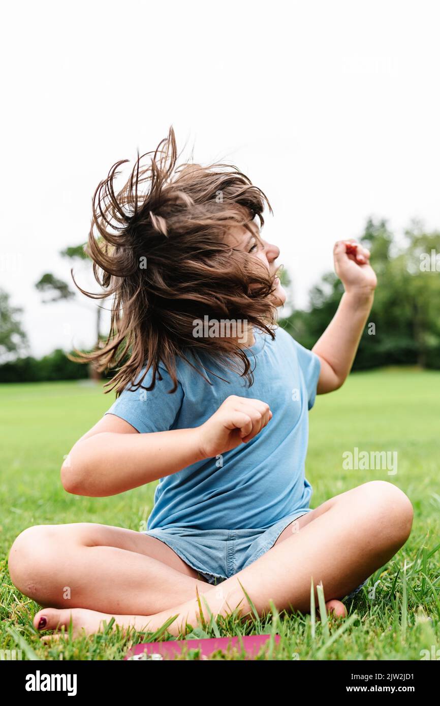 Cheerful barefoot child in casual clothes smiling and shaking hair while sitting cross legged on grassy lawn on summer weekend day in park Stock Photo
