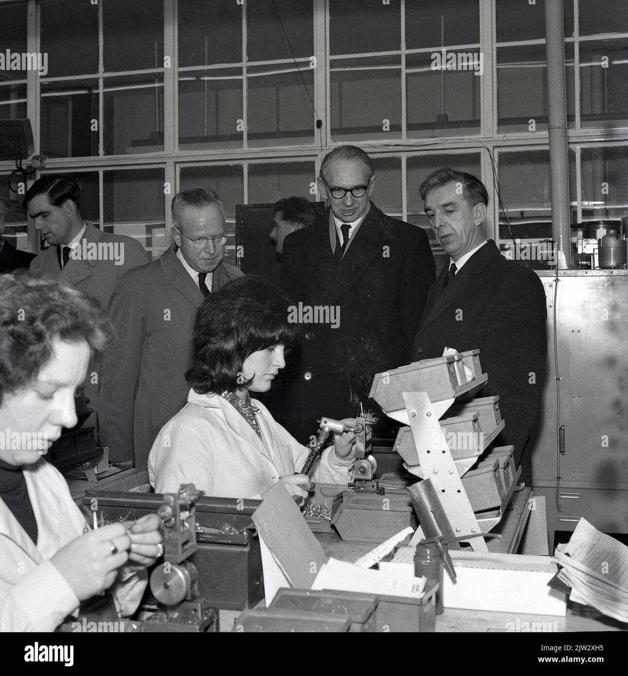 1960s, historical, visiting company executives on the shop floor in a light engineering factory, watching female employees in white-coats sitting working, Fife, Scotland, UK. The rise of Silicon Glen started in 1943 when Ferranti, an electrical engineering and equipment firm, moved north of the border from from Manchester. Gradually through the 50s & 60s, overseas companies started a production facility there, including IBM and the Scottish electronic sector by1990, known as Silicon Glen was at it’s peak. Stock Photo