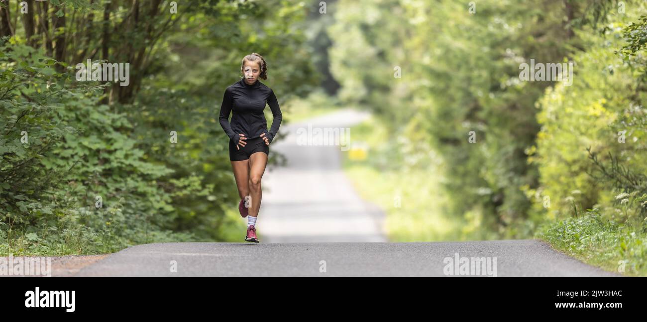Young female athlete runs up the hill on an asphalt road in the nature. Stock Photo