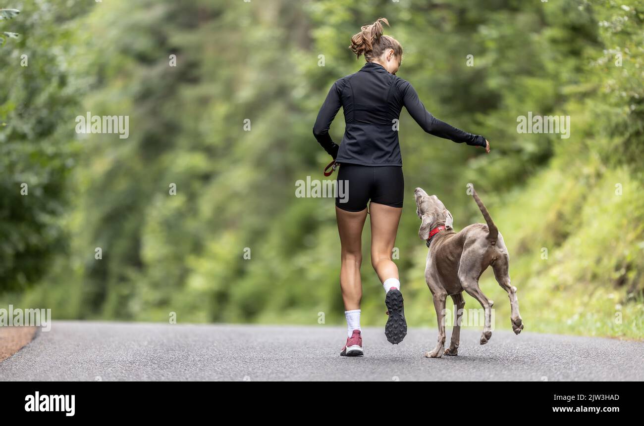 Picture from behind of a girl running in the nature with her dog jumping on her side. Stock Photo