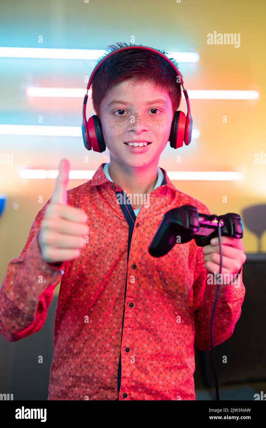 portrait shot of young Teenager kid with headphones holding gamepad by showing thumbs up by looking at camera at home - concept of live streamer and Stock Photo