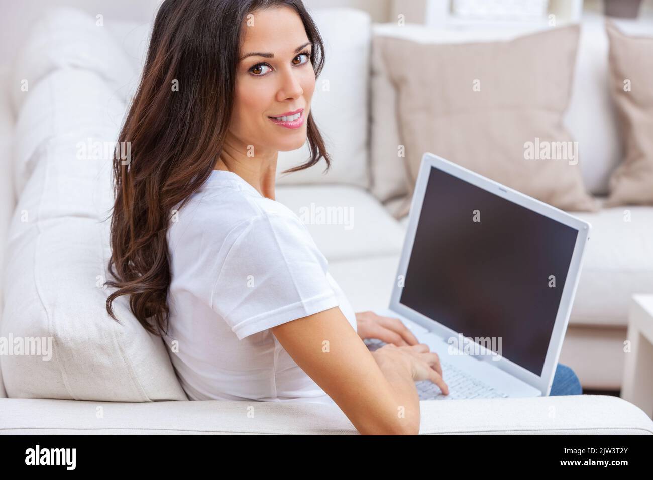 Beautiful girl female young woman at home sitting on sofa or settee using her laptop computer to work from home, on line shopping or social media Stock Photo
