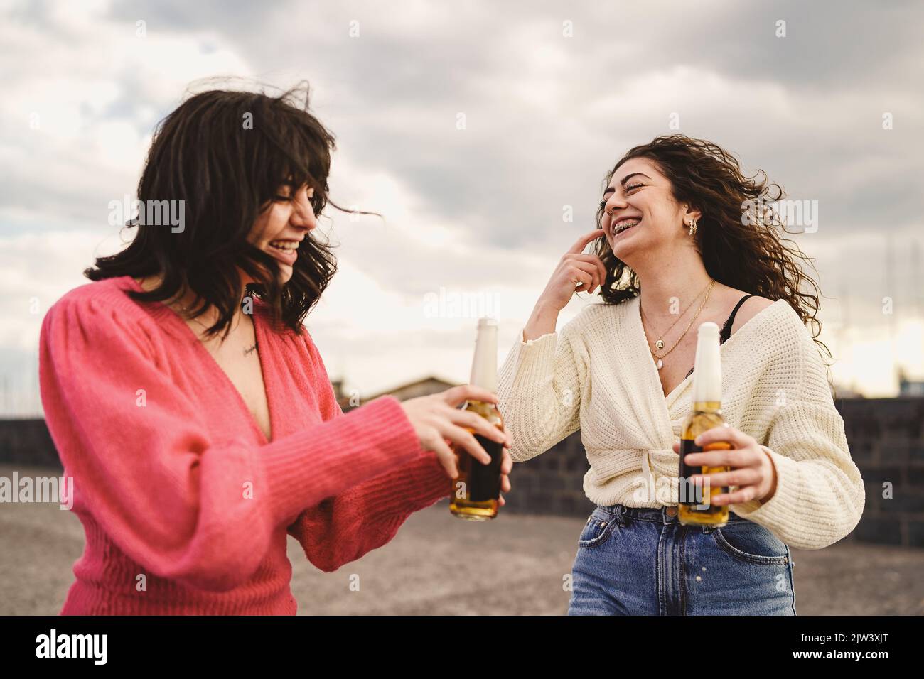 Two girlfriends laugh together with beers whose foam comes out of the bottle - young women drinking beers and having fun together outdoors - people li Stock Photo