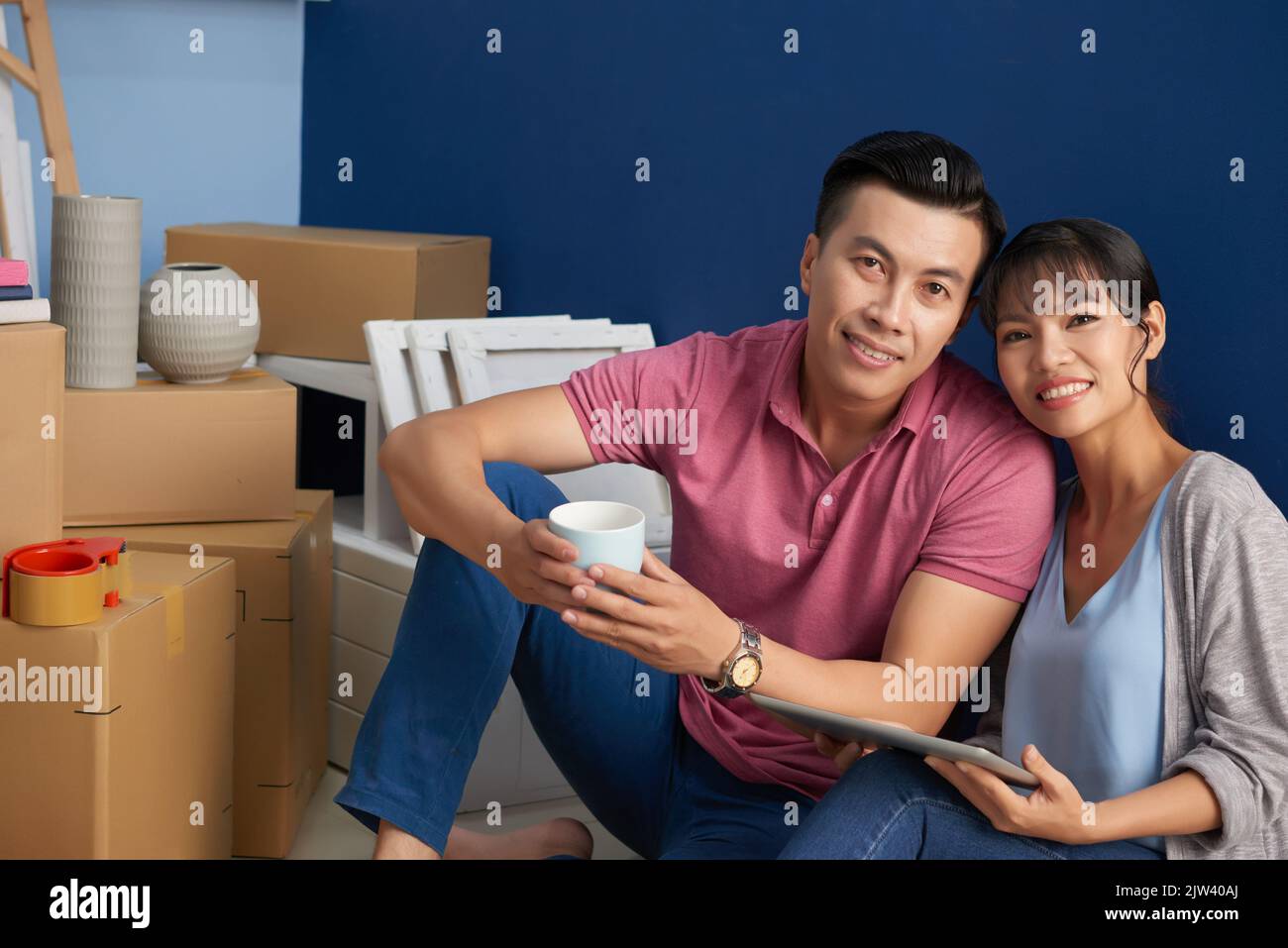 Portrait of young Asian couple smiling at camera happily while sitting in new flat near packed carton boxes and household stuff Stock Photo