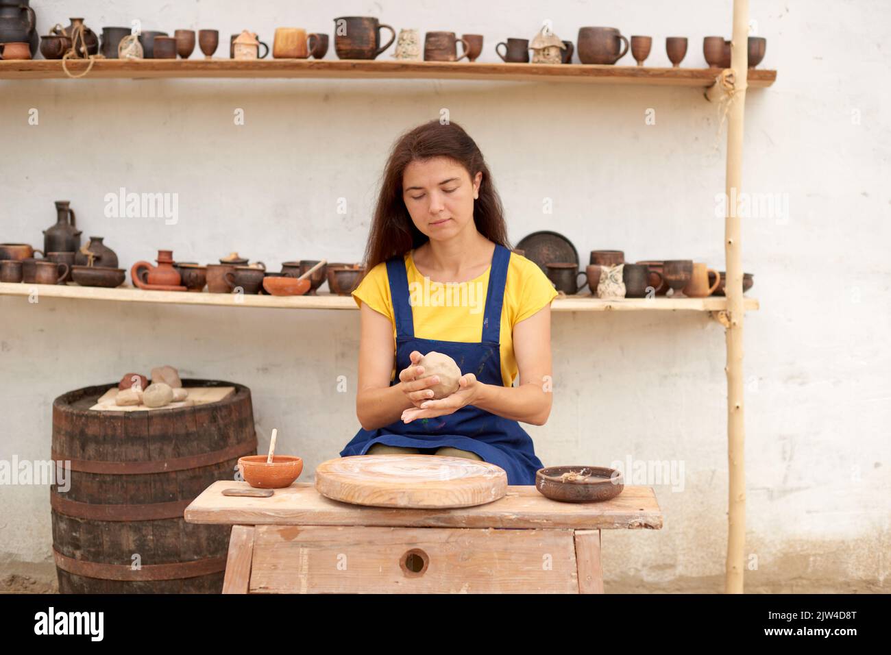 woman in blue apron and yellow T-shirt works on mechanical potter's wheel against background of shelves of clay products Stock Photo