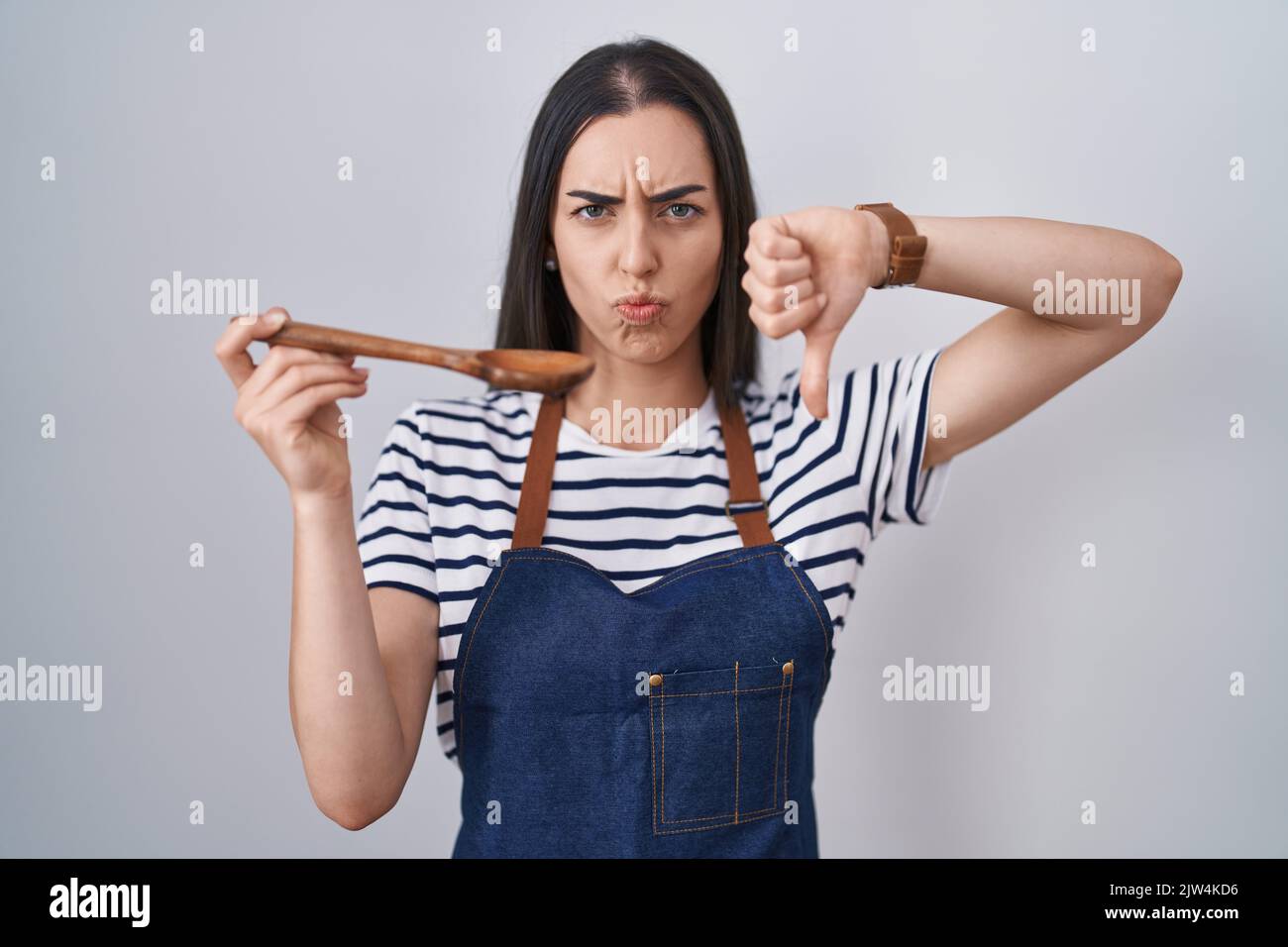 Young brunette woman wearing apron tasting food holding wooden spoon ...