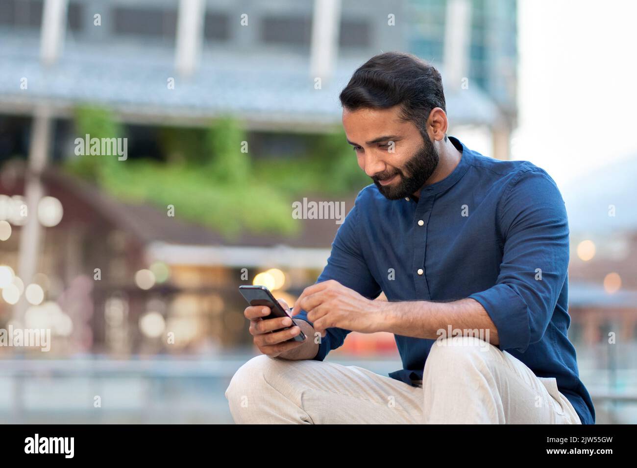 Smiling indian businessman using smartphone sitting on urban city street. Stock Photo