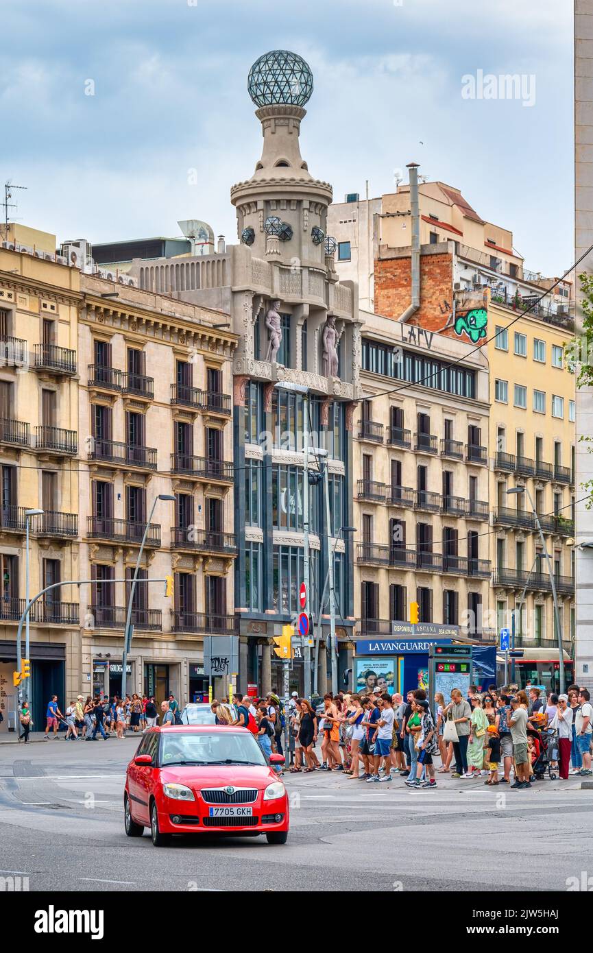 A crowd of people waits to cross the street in the Plaza de Catalonia. The famous architecture of buildings is seen in the background. Stock Photo