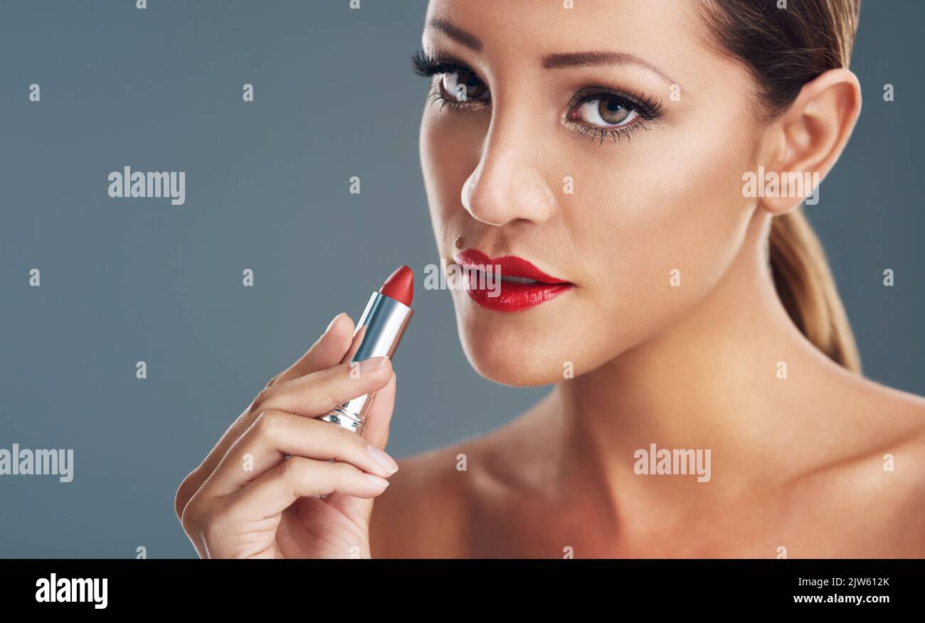 Vamp up your makeup routine with red lipstick. Studio portrait of a beautiful young woman putting on red lipstick against a grey background. Stock Photo