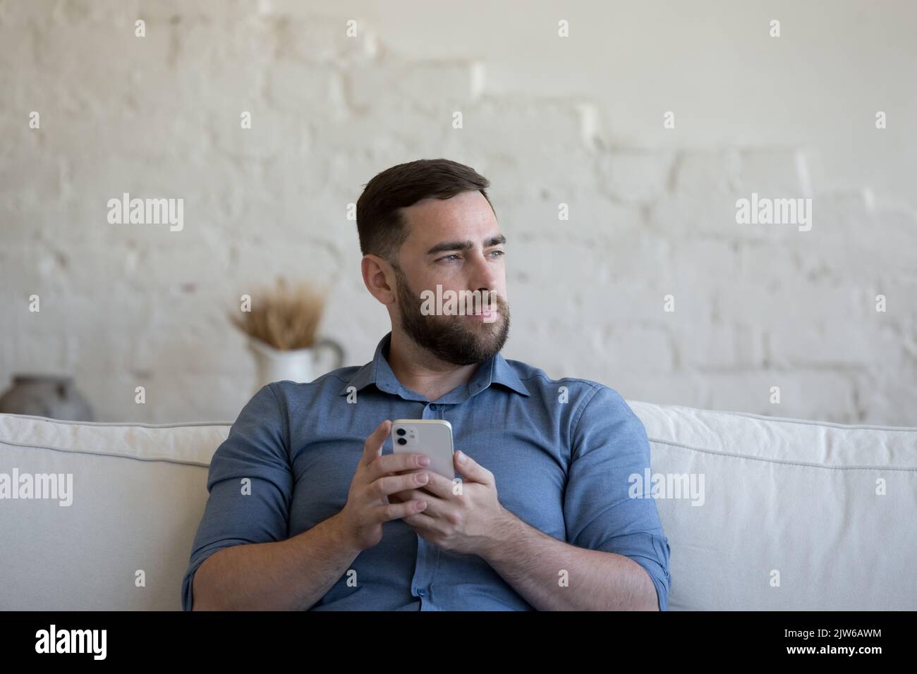 Pensive millennial man holds smartphone seated on sofa Stock Photo