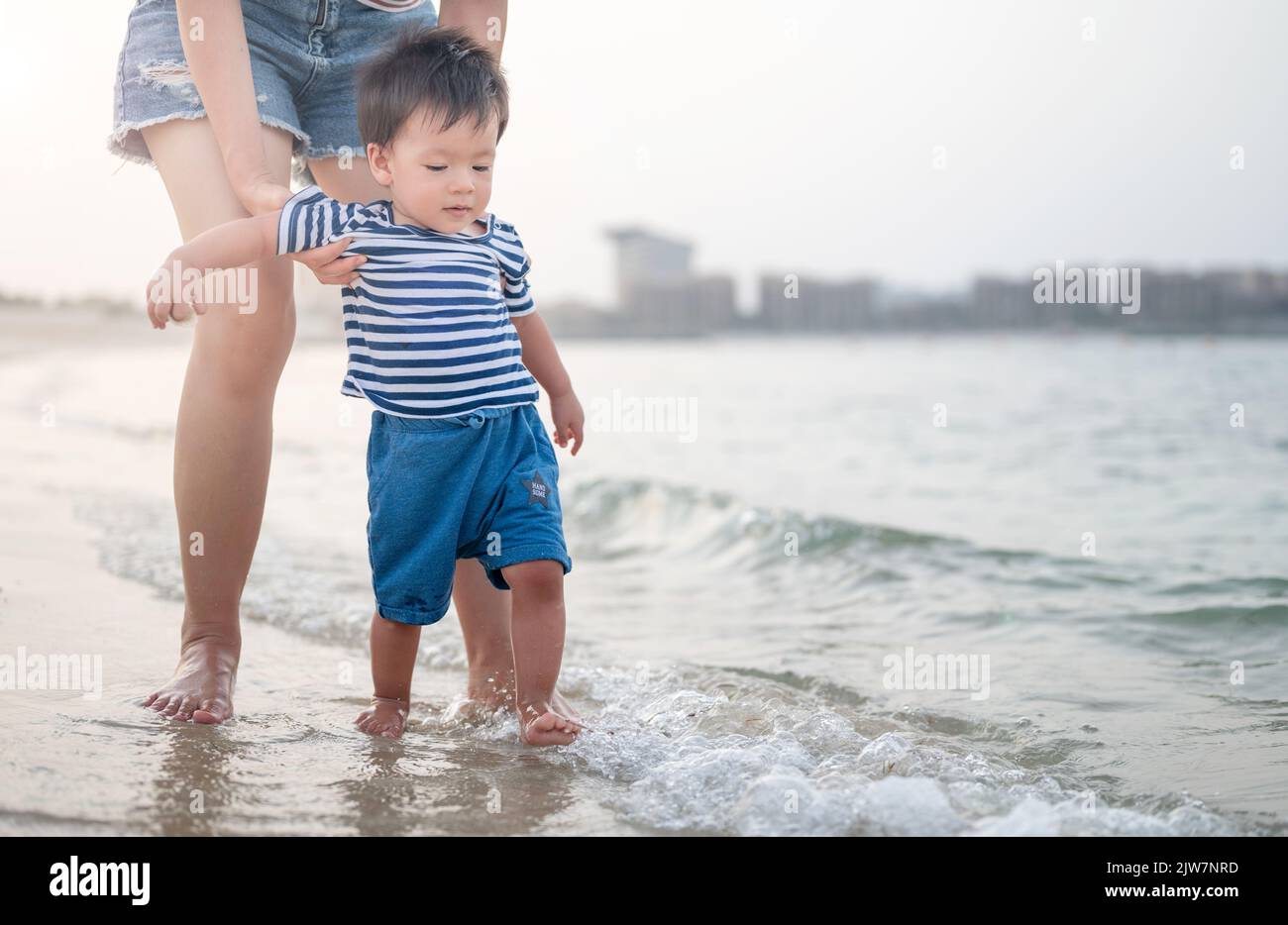 Adorable baby boy making his first steps on the beach by the seaside with his mother. One year old Infant learning how to walk on a summer vacation Stock Photo