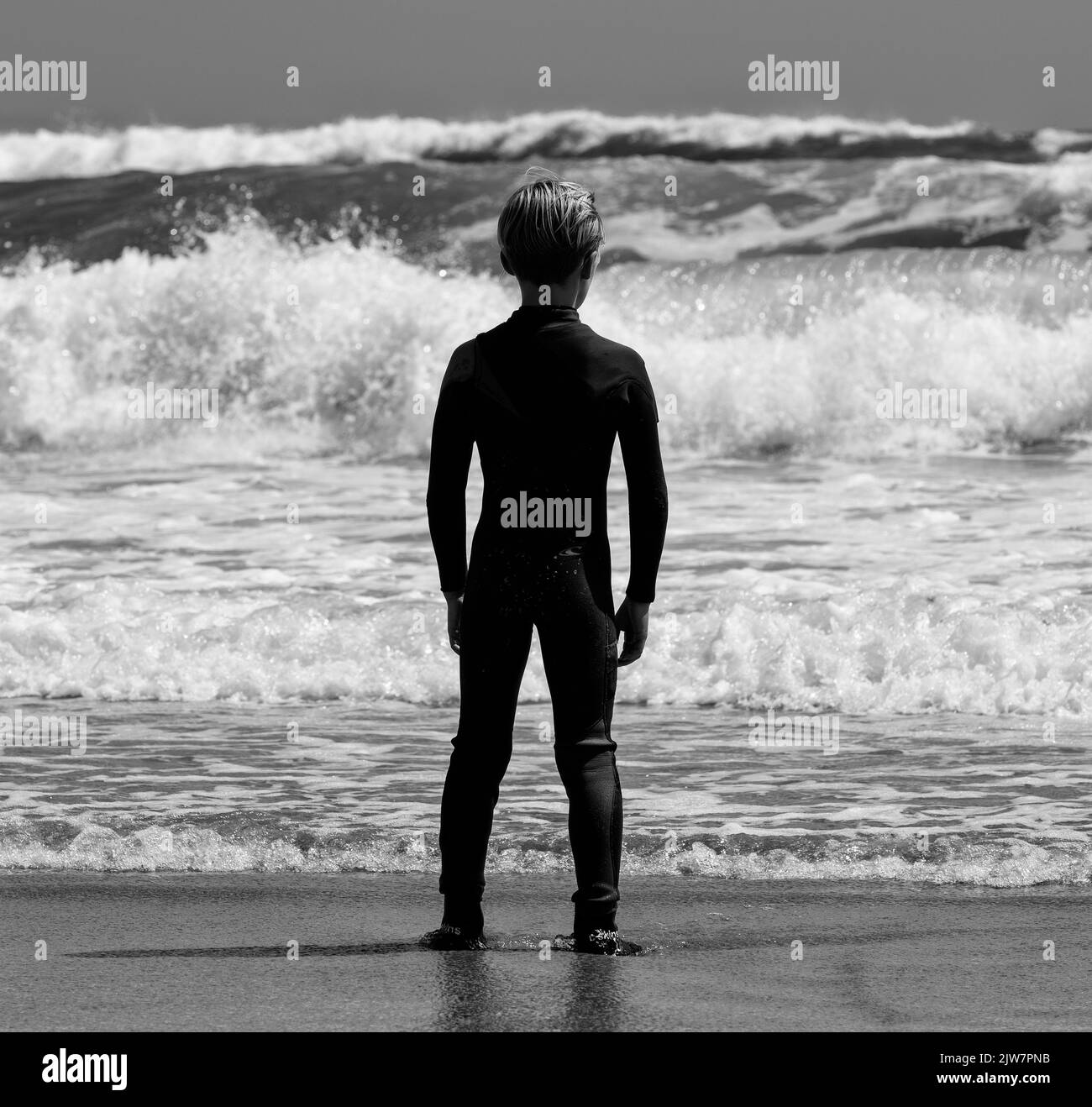 young lad Surfer enjoying the waves at Gwithian sands beach Cornwall. Black and white. Taking time to look out to sea. Stock Photo