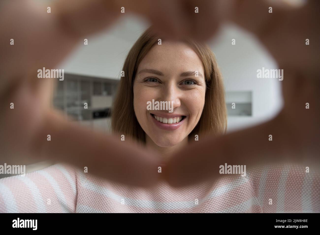 Happy adult woman looking at camera through finger heart Stock Photo