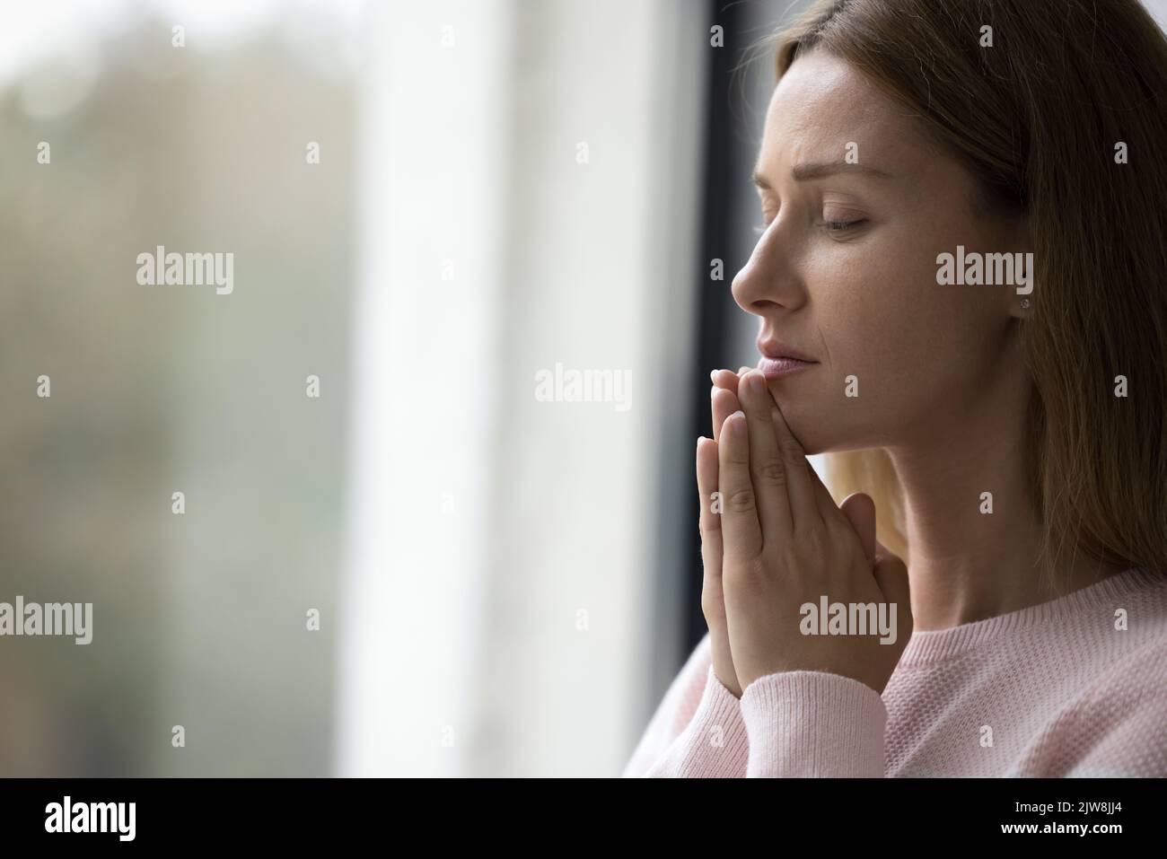 Serious calm pretty adult woman standing at window indoors Stock Photo