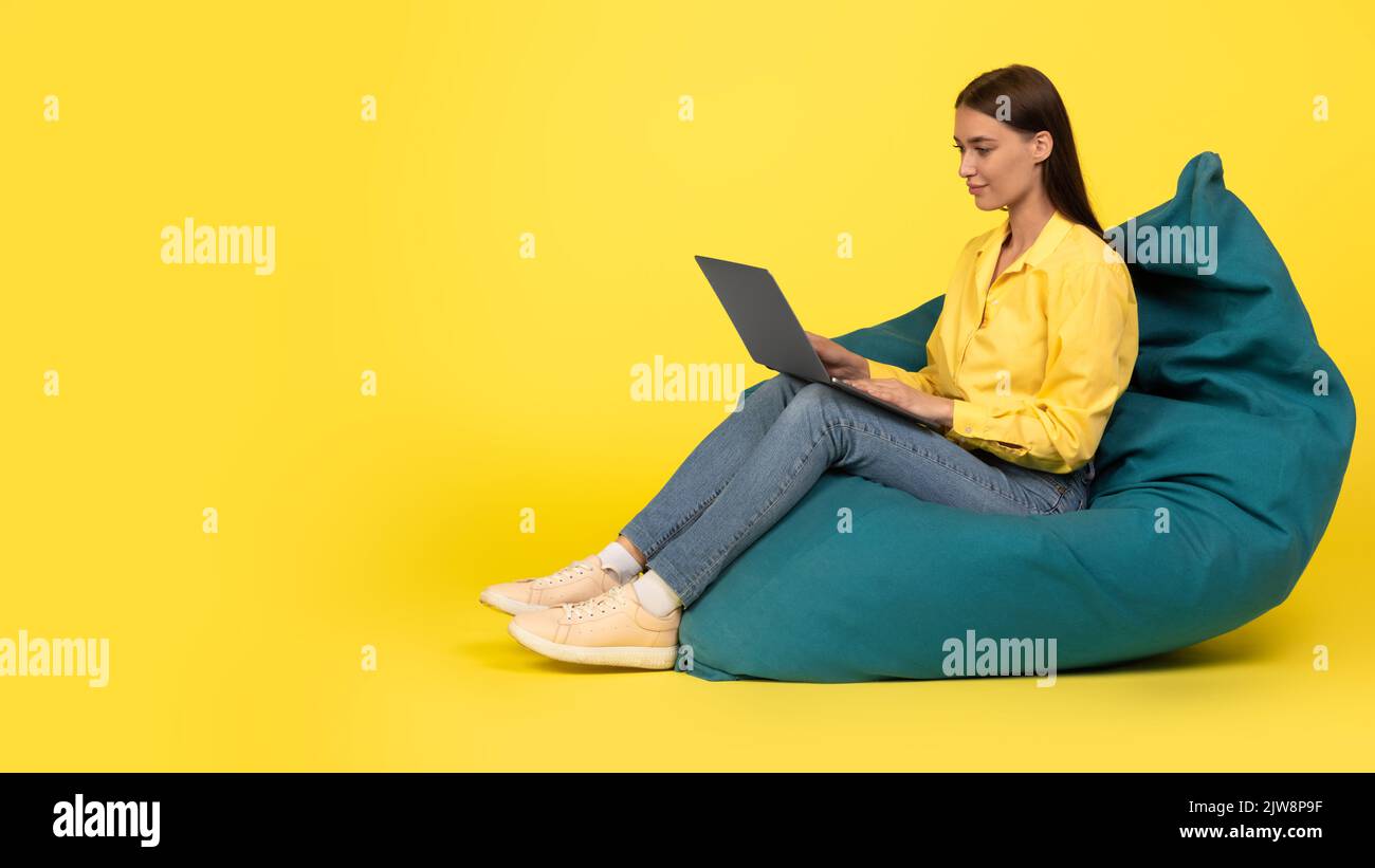 Female Using Laptop Working Sitting In Beanbag Chair, Yellow Background Stock Photo