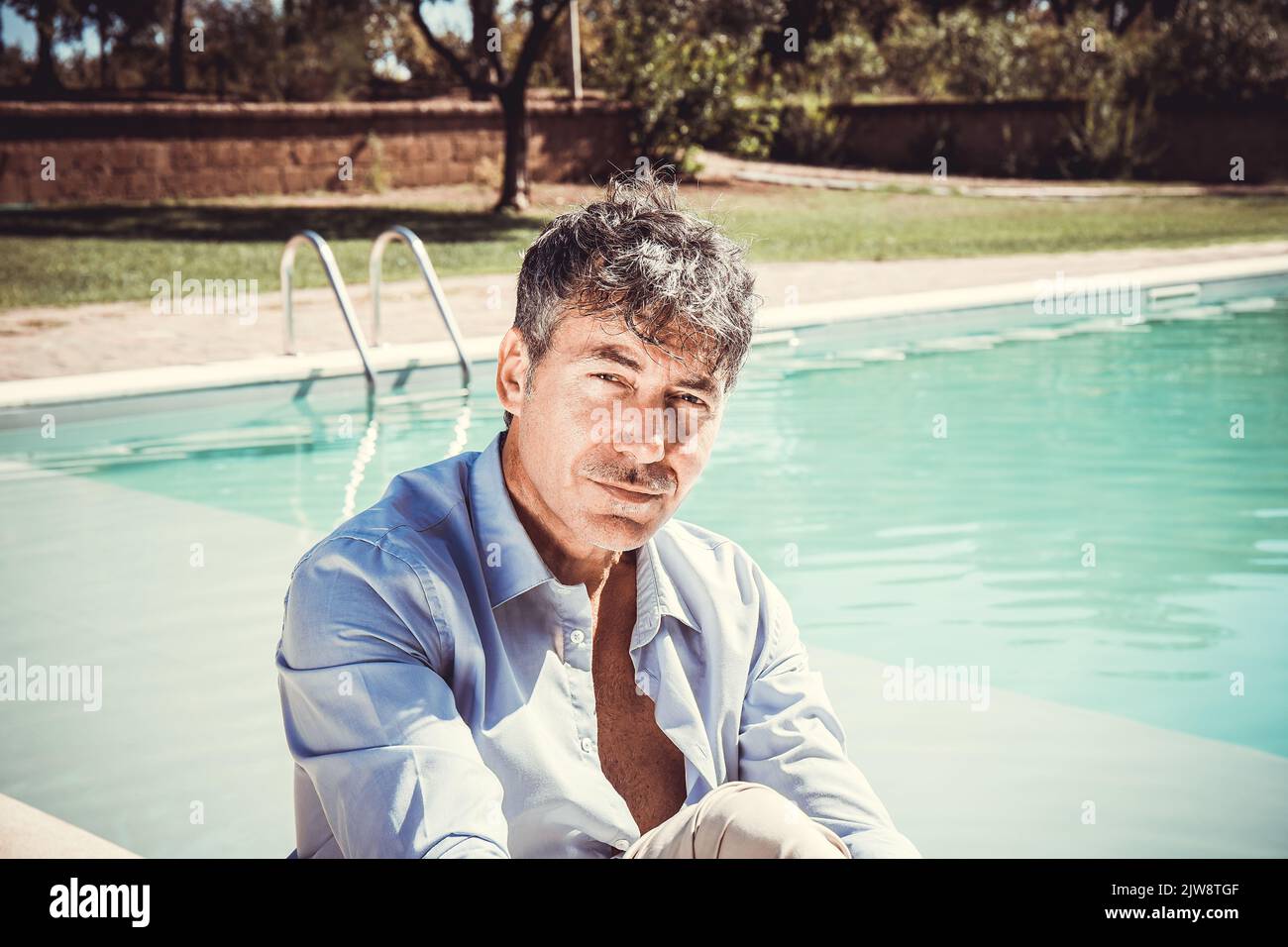 The young man is wearing sunglasses, white shirt and beige pants and is sitting by the pool. A handsome businessman takes a moment to relax by the poo Stock Photo
