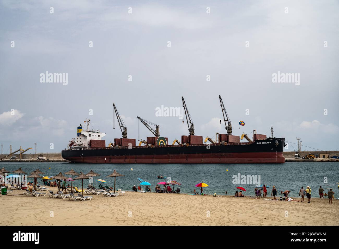 Tourists and locals enjoy a summer day at Garrucha Beach, in the province of Almeria, south of Spain, while a cargo ship is at the port Stock Photo