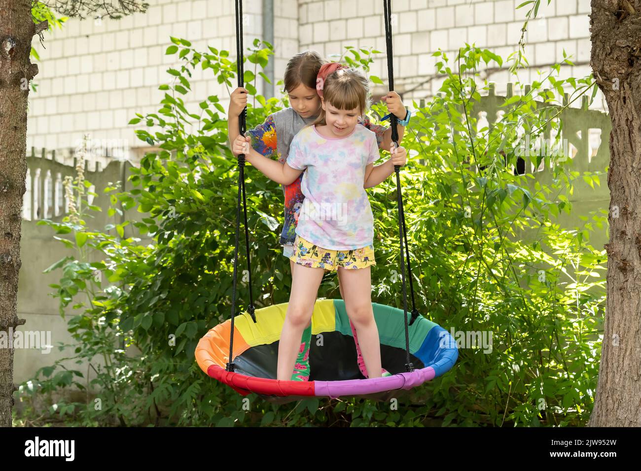 Two girls, children standing together on a swing in the garden, having fun outdoors, smiling. Fun summer activities, sisters, siblings, friends bondin Stock Photo