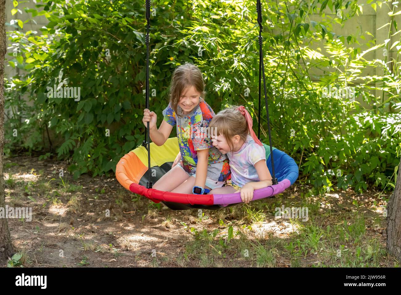 Two children, girls, kids sitting on an outdoor swing in the garden laughing together, having fun outdoors. Outside activities, laughter, real people Stock Photo
