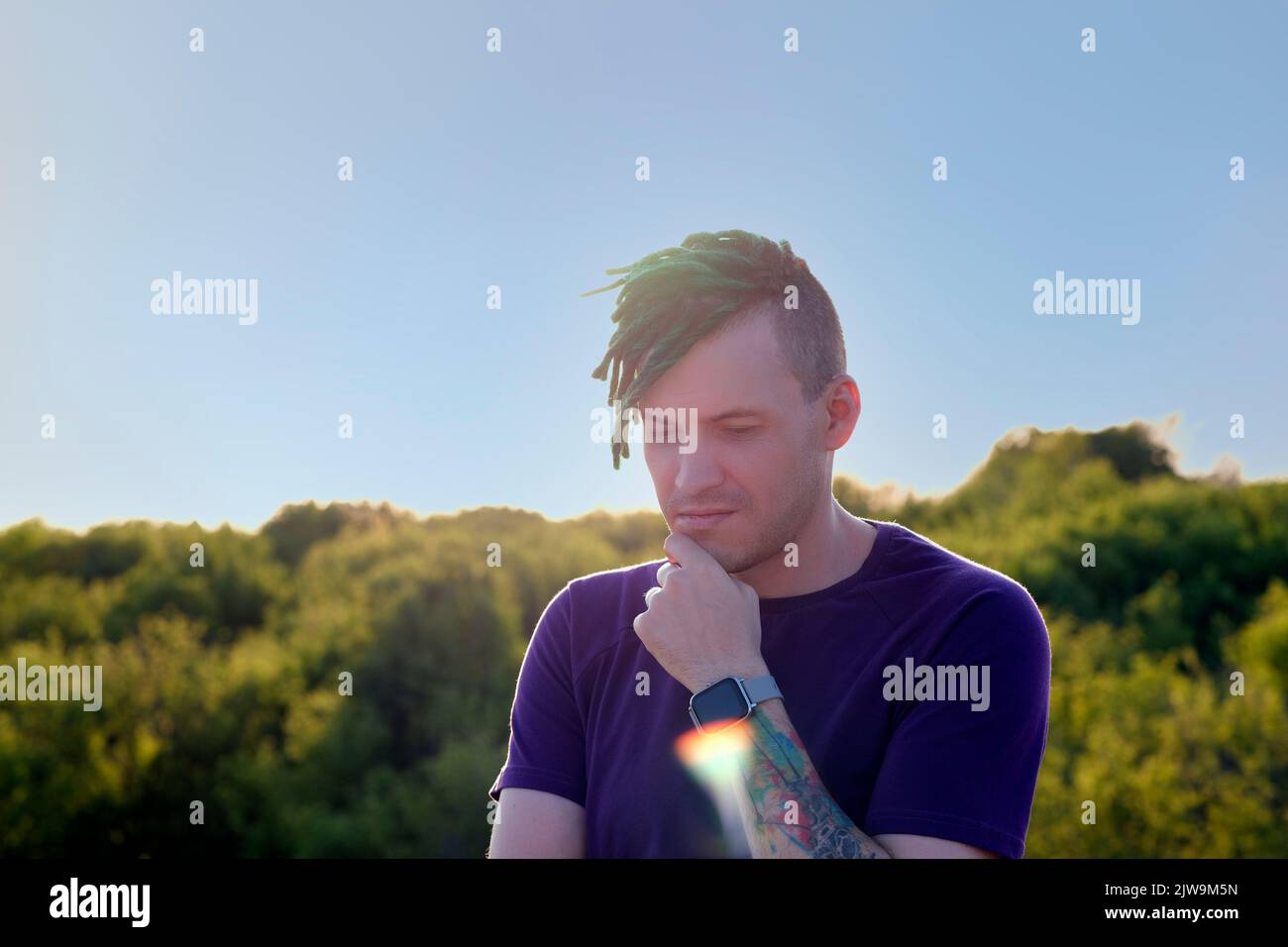 Young thoughtful man with green dreadlocks in sunglasses looking away, scratching his chin with finger, standing on hill on sunset Stock Photo