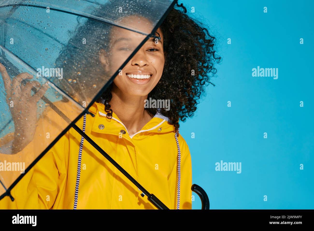 Portrait of smiling African woman with afro-hairstyle in yellow raincoat isolated over blue background with wet transparent umbrella Stock Photo