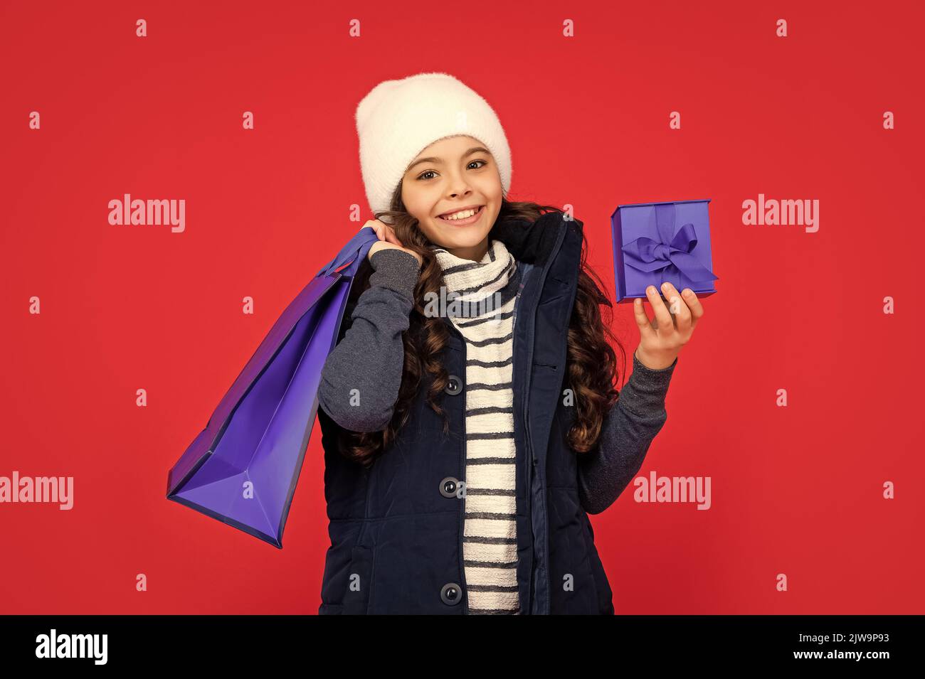 cheerful kid in puffer jacket and hat. teen girl after shopping on red background. child holding shopping bag and gift box. winter sales. Stock Photo