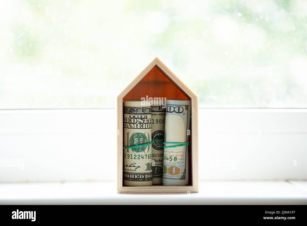 A small wooden house with hundred dollar bills inside on the window of the house, buying a house, finance and real estate Stock Photo