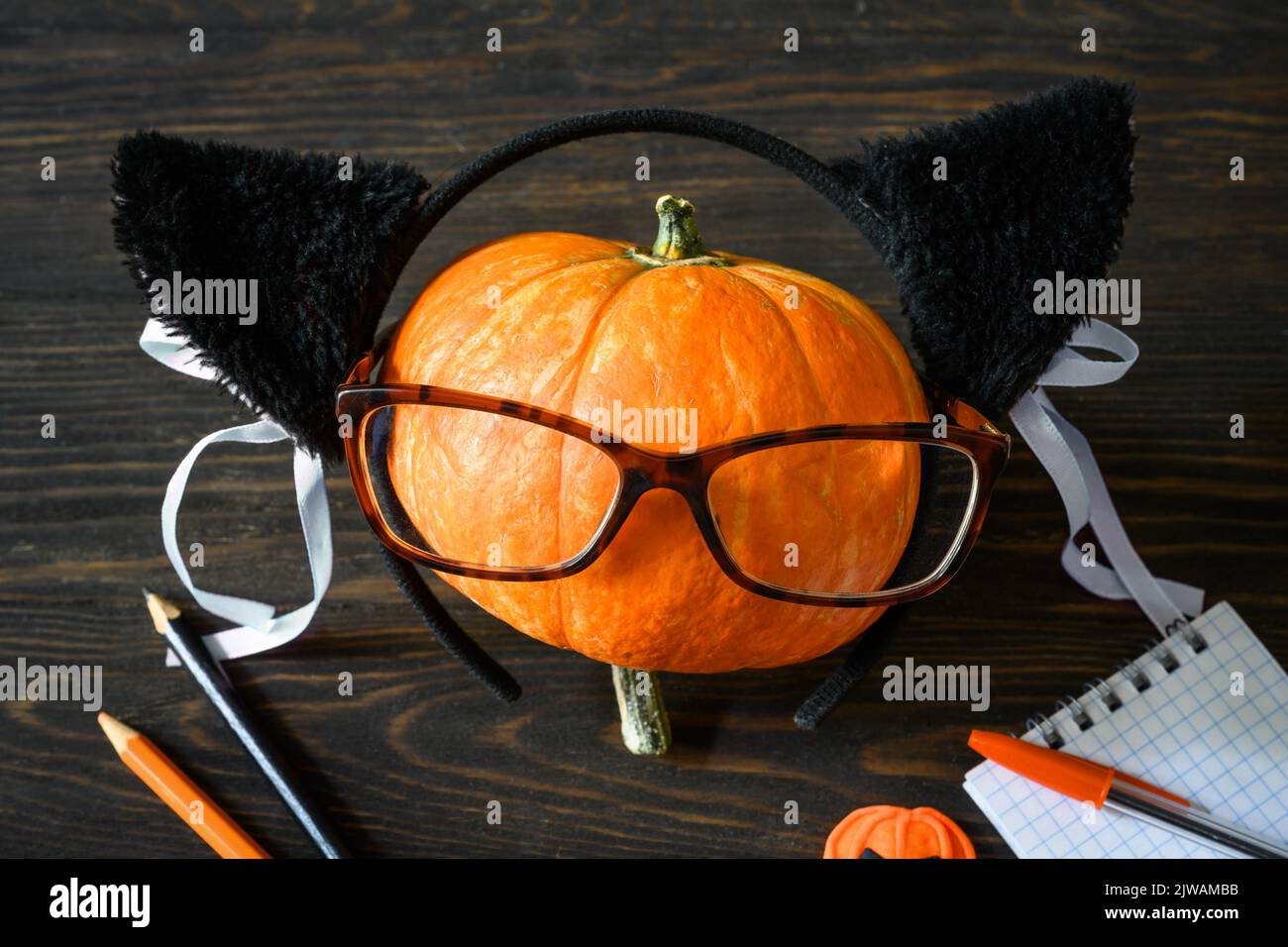 Halloween still life, happy pumpkin with glasses on wooden table, school supplies on Halloween. Orange pumpkin, notepad, pens, pencil on dark wood. Co Stock Photo