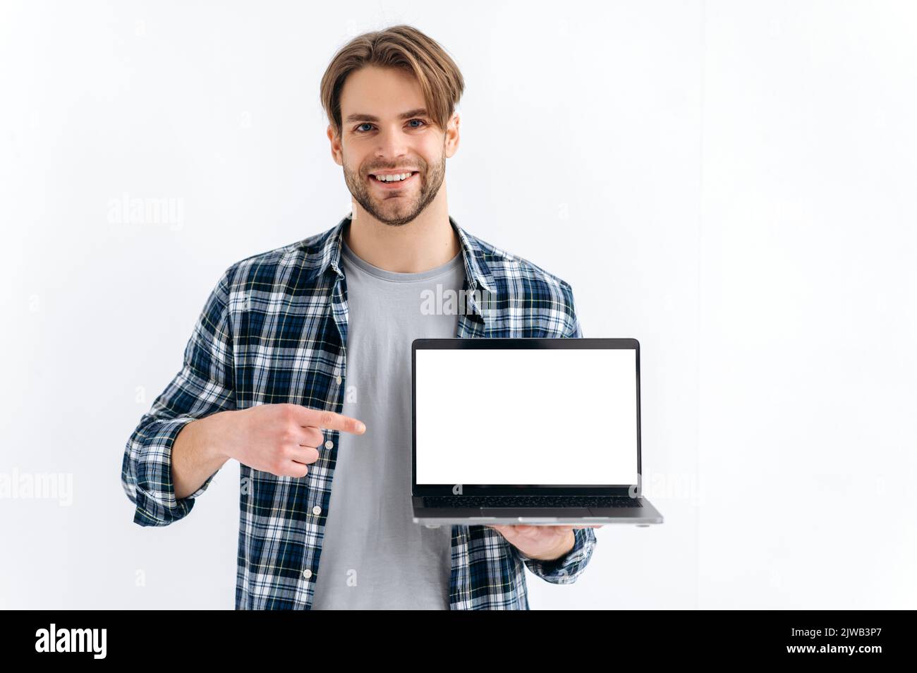 Stylish handsome confident caucasian man, standing on a white isolated background, holds an open laptop, showing empty white mock-up screen for advertising or presentation, points finger at it, smile Stock Photo