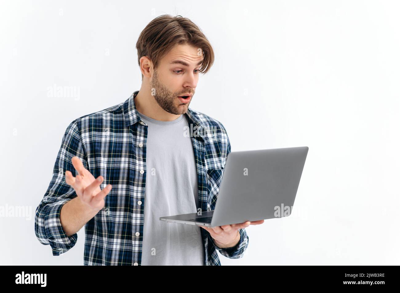 Shocked confused puzzled caucasian stylish young man, standing on a white isolated background, holds an open laptop, surprised looks at the screen, see unexpected news, message, gesturing with hand Stock Photo