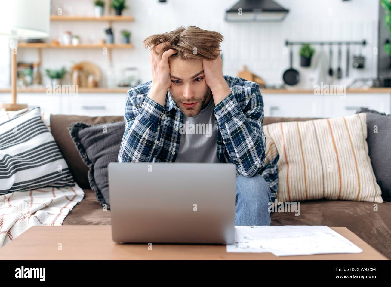 Confused upset caucasian young stylish man, sitting on sofa in living room, working online, frustrated looking at laptop screen, failed project, failed deal, holding hands on head, despair emotions Stock Photo