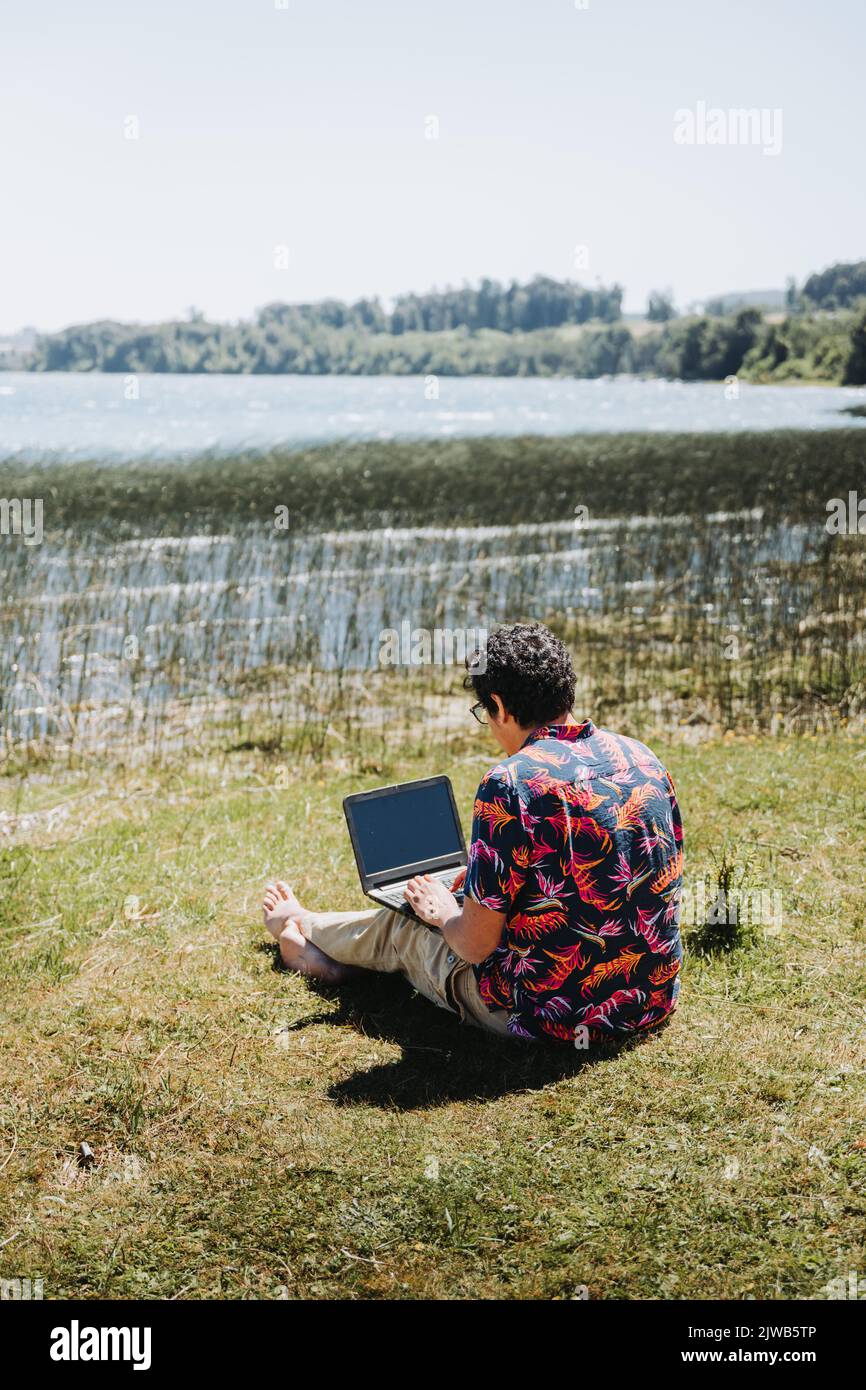 Rear view young latin man doing remote online work from the lake. Teleworking concept. Digital nomad Stock Photo