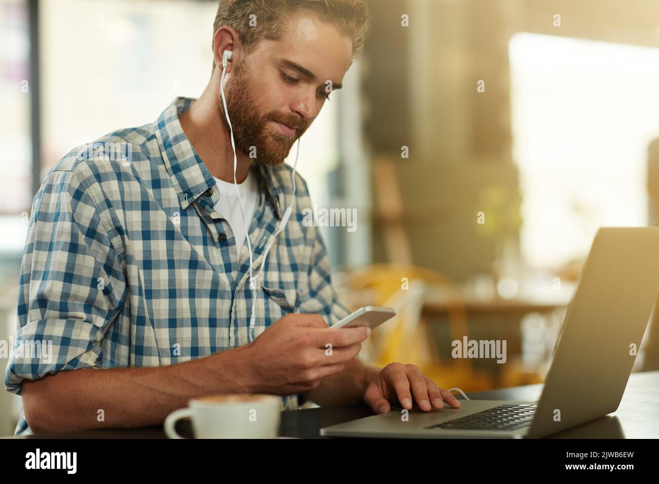 Plans seem to be going smoothly. a young man with earphones using a cellphone and laptop in a cafe. Stock Photo