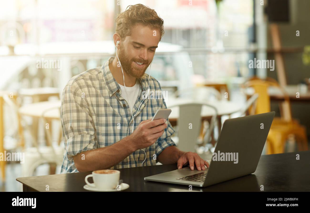 Staying connected even outside of the office. a young man with earphones using a cellphone and laptop in a cafe. Stock Photo