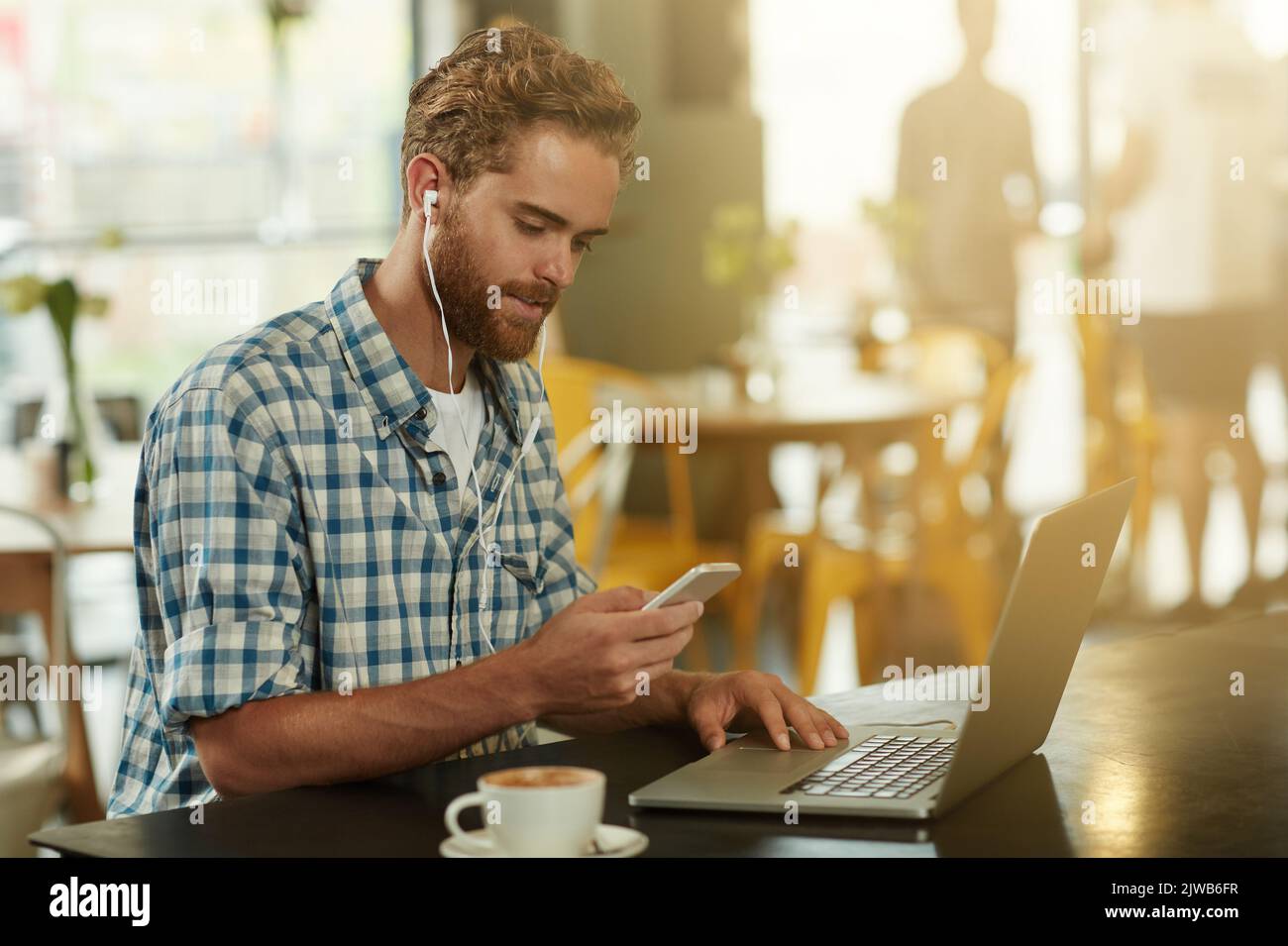 Relaxed and productive. a young man with earphones using a cellphone and laptop in a cafe. Stock Photo
