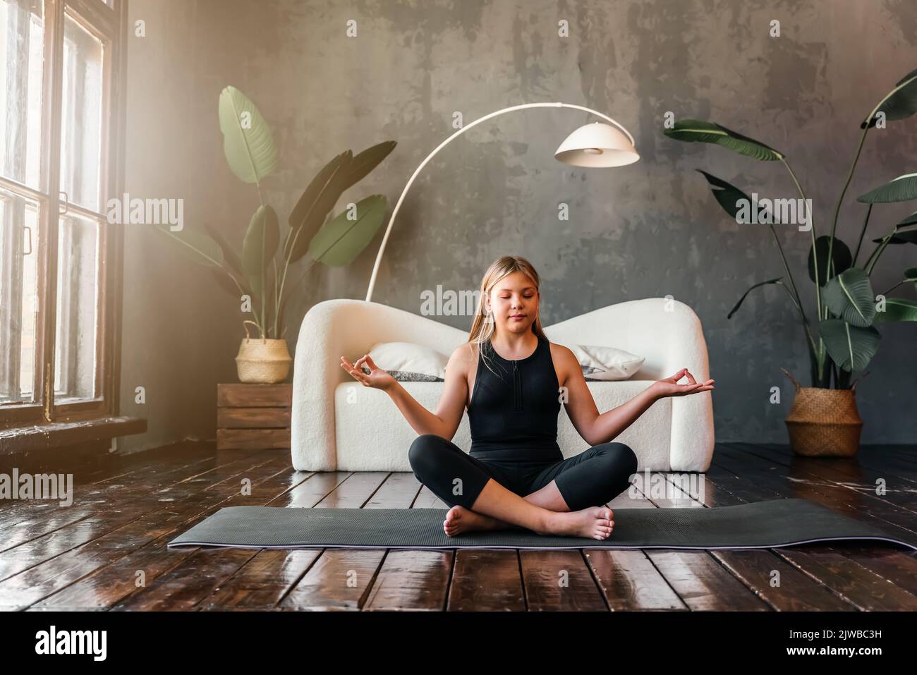 Young attractive girl practicing yoga, sitting in Half Lotus exercise, Ardha Padmasana pose, exercising, meditation session, home interior. High quali Stock Photo