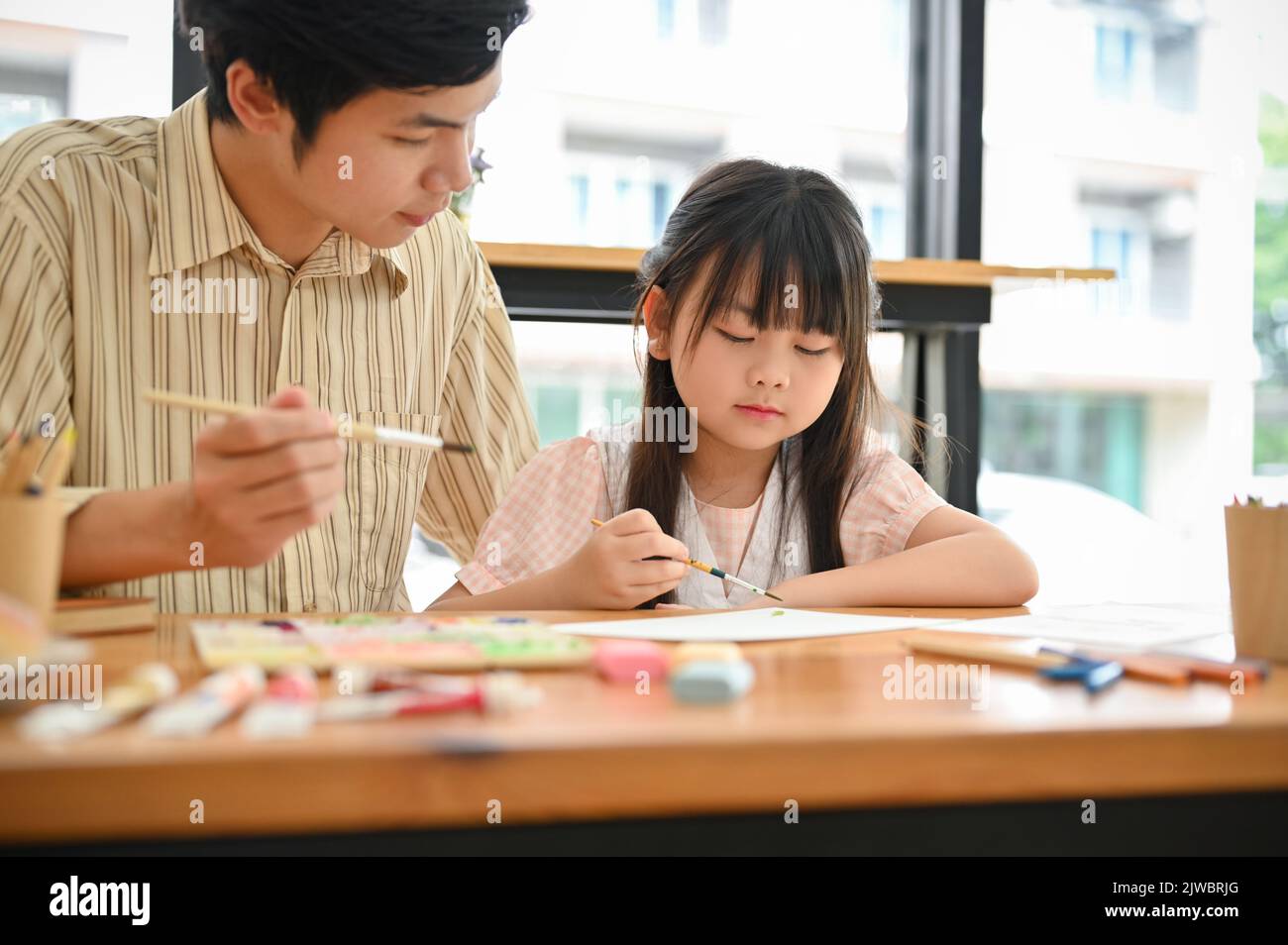 A cute little Asian girl is concentrating painting picture with a watercolor, learning art with her private teacher. Kids' leisure concept Stock Photo