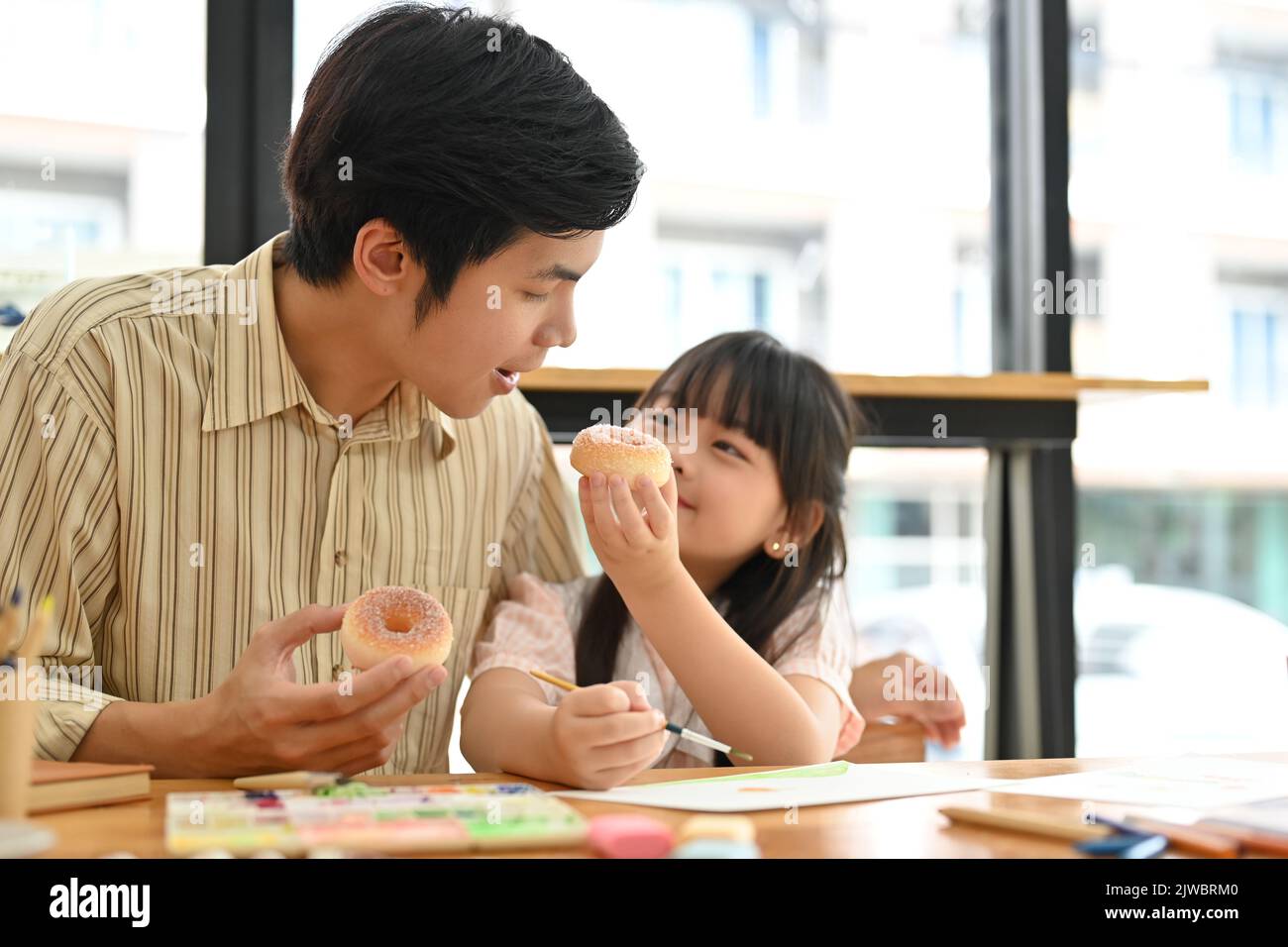 A kind and lovely young Asian girl shares a doughnut with her dad in the cafe. having a great time together, enjoying activity, happy family moment. Stock Photo