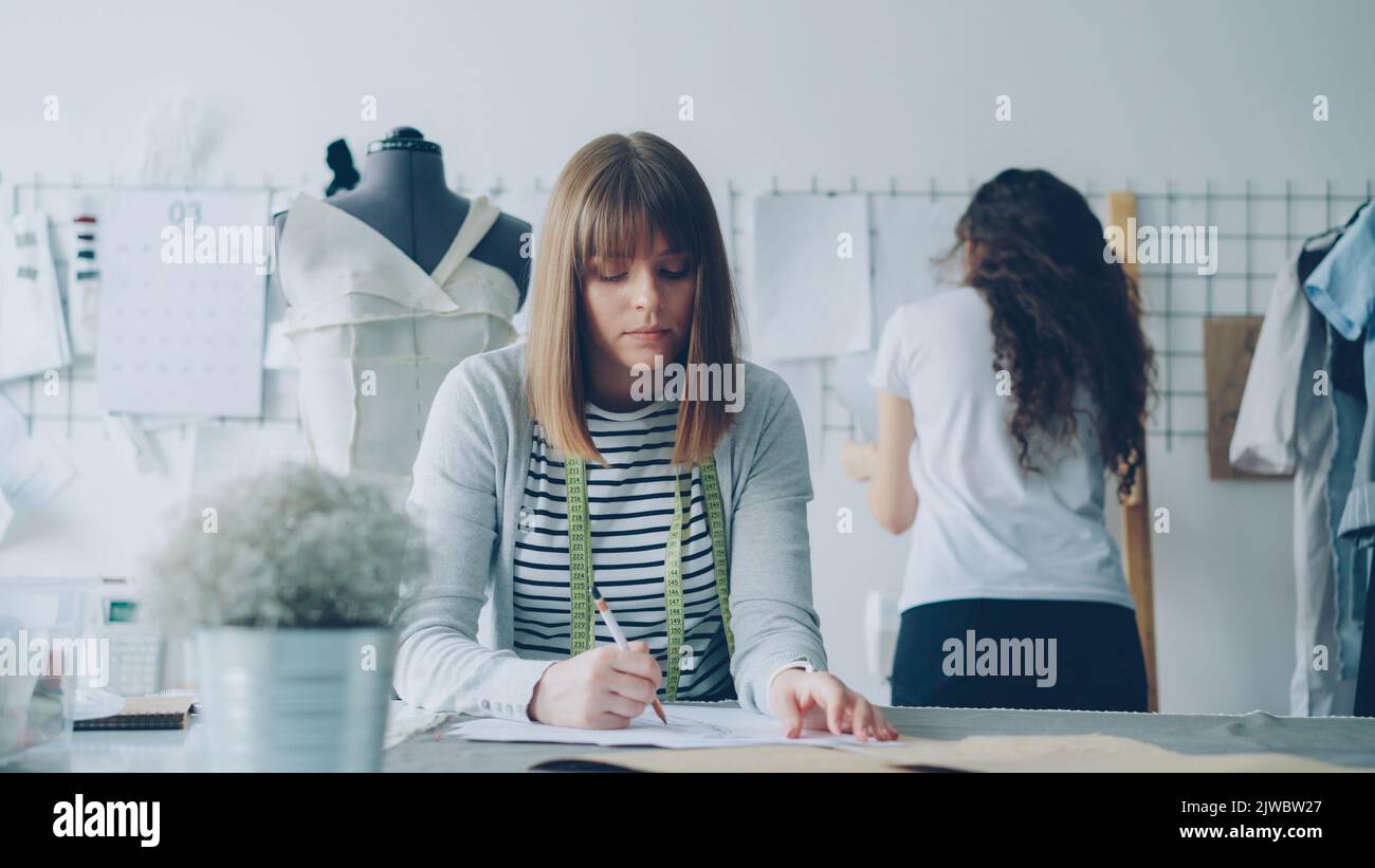 Young attractive clothing designer is busy drawing sketch with pencil while her colleague is watching pictures pinned on wall in design studio. Creative thinking concept. Stock Photo