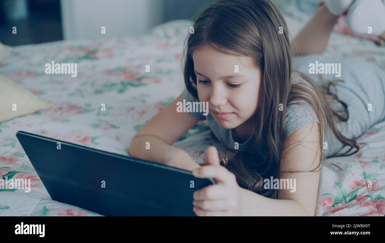 Cute little girl using digital tablet and smiling while lying in bed. Child wathcing cartoon movie on portable device and laughing at home Stock Photo