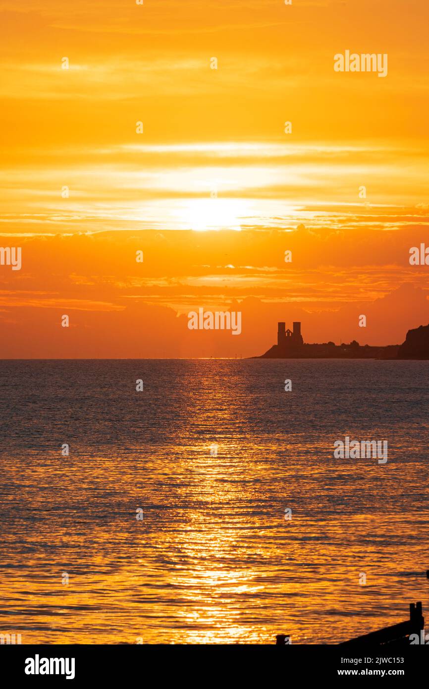 Sunrise over the sea on the Kent coast at Reculver with the twin towers of the 12th century church silhouetted against an orange sky. The ruined church is a well-known landmark Stock Photo