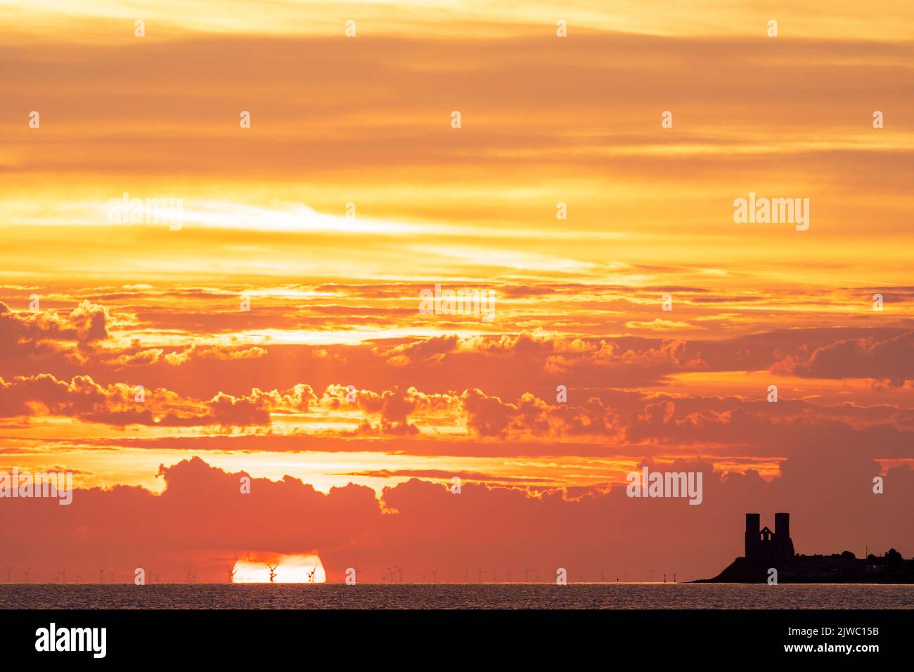 Sunrise over the sea on the Kent coast at Reculver with the twin towers of the 12th century church silhouetted against an orange sky. The ruined church is a well-known landmark Stock Photo