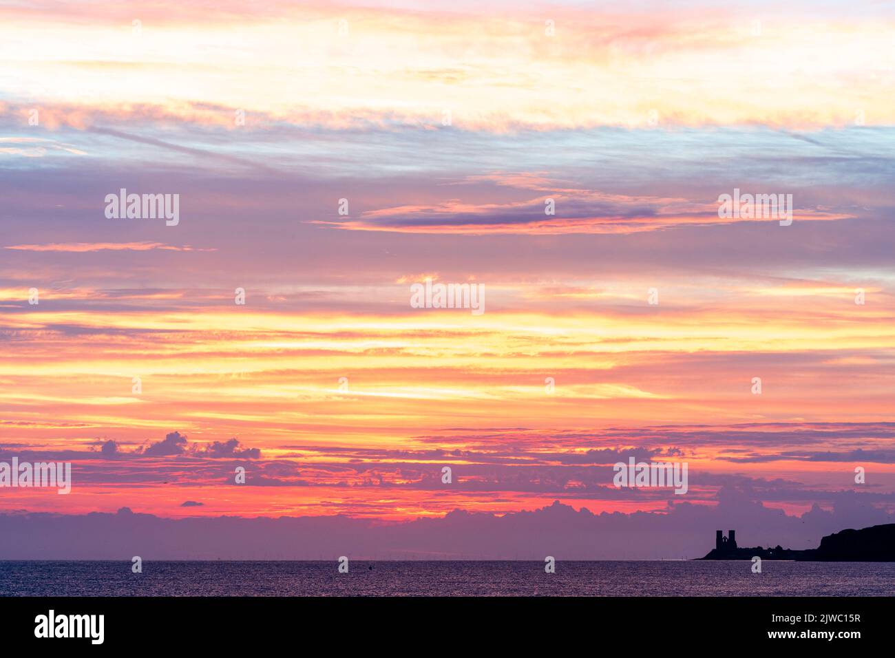 Scattered cloud in the dawn sky over the sea at the Kent coast at Reculver. The multi-coloured clouds are lit from underneath by the rising sun still below the horizon. In the distance is the twin towers of the ruined 12th century Reculver church. Stock Photo