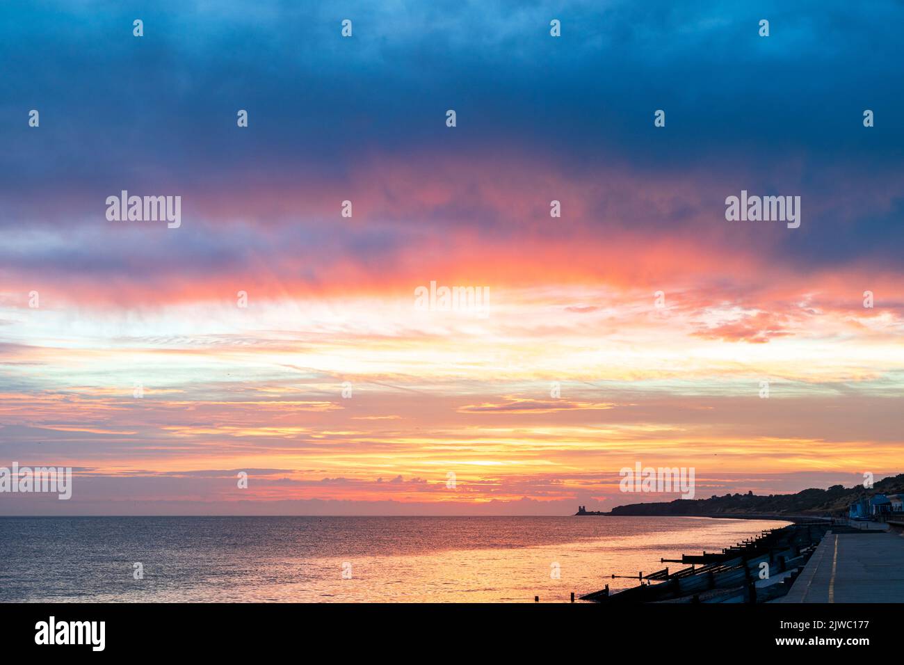 The dawn sky over the sea on the Kent coast at Reculver with the twin towers of the 12th century church silhouetted in the distance against an orange sky. In the foreground the silhouetted breakwaters, groynes, and beach at Henre Bay. Thick cloud overhead underlit by orange light. Stock Photo
