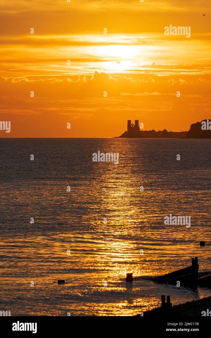 Sunrise over the twin towers of the 12th century church at Reculver and the sea at Herne Bay. Bright orange sky with a ray of reflected light across the sea silhouetting the groynes, breakwaters on the Henre Bay seafront. Stock Photo